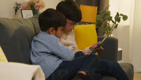 Two-Young-Boys-Sitting-On-Sofa-At-Home-Playing-Games-Or-Streaming-Onto-Digital-Tablet-Together-4